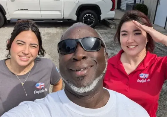 Three smiling people posing outdoors in front of a white vehicle, with the center person taking the selfie.