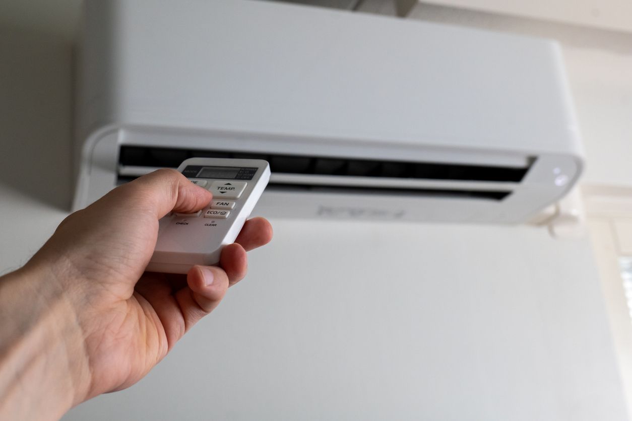 Hand holding a remote pointed at a wall-mounted air conditioner in a bright room