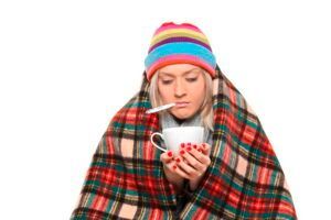 Person wrapped in a plaid blanket, wearing a colorful beanie, holding a steaming mug and looking unwell