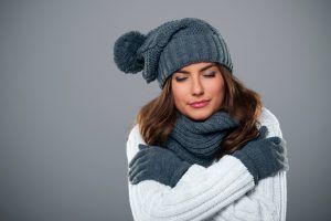 Person in a blue knit hat and scarf, hugging themselves against a gray background.