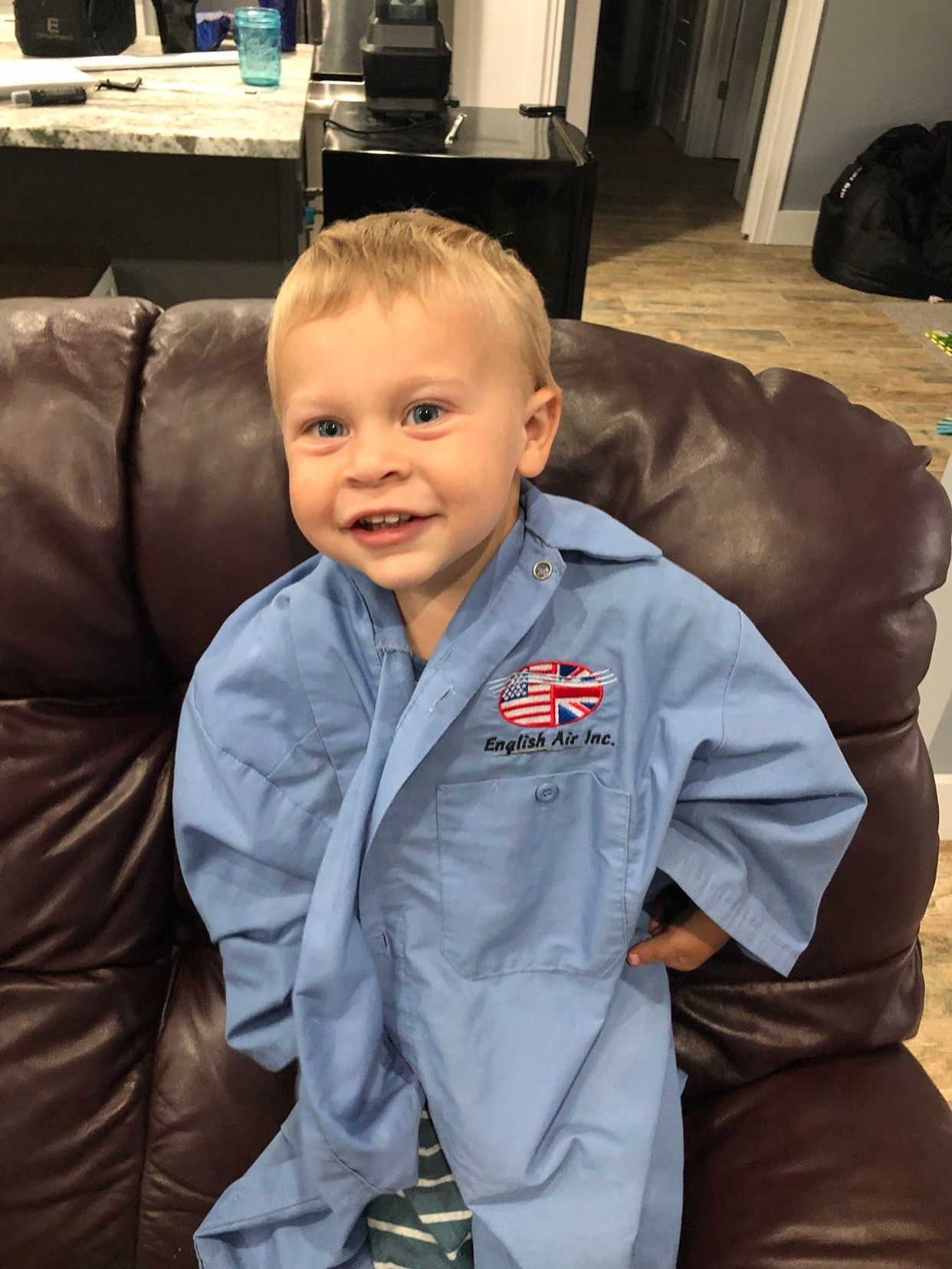Smiling toddler in a blue jumpsuit standing on a brown leather couch indoors.