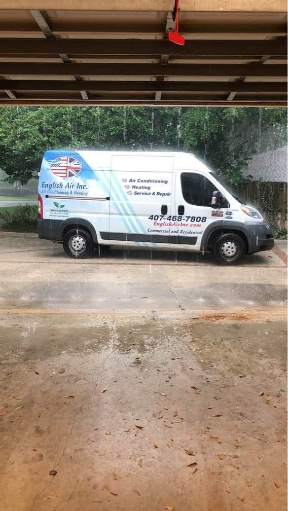 White delivery van parked under a roof, with blue graphics and text on the side, near wet pavement.