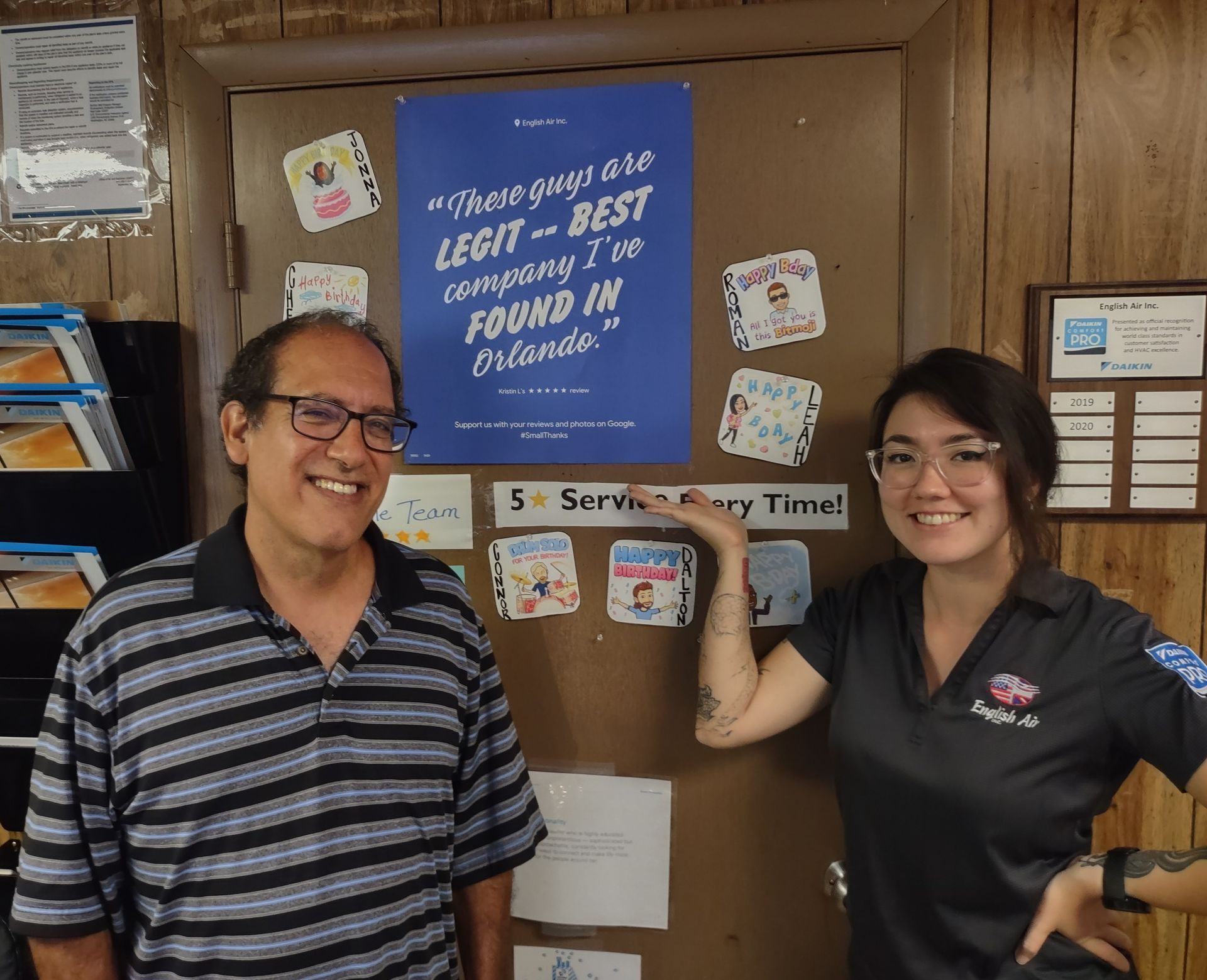 Two smiling people stand beside a blue sign on a cardboard backdrop in a room.