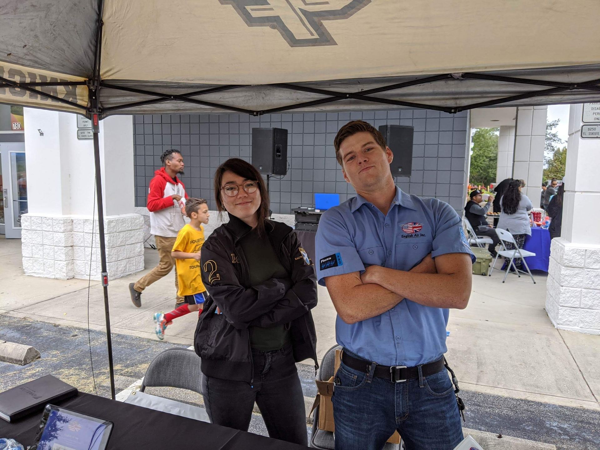 Two people standing under a canopy at an outdoor event, arms crossed, with people and tents in the background