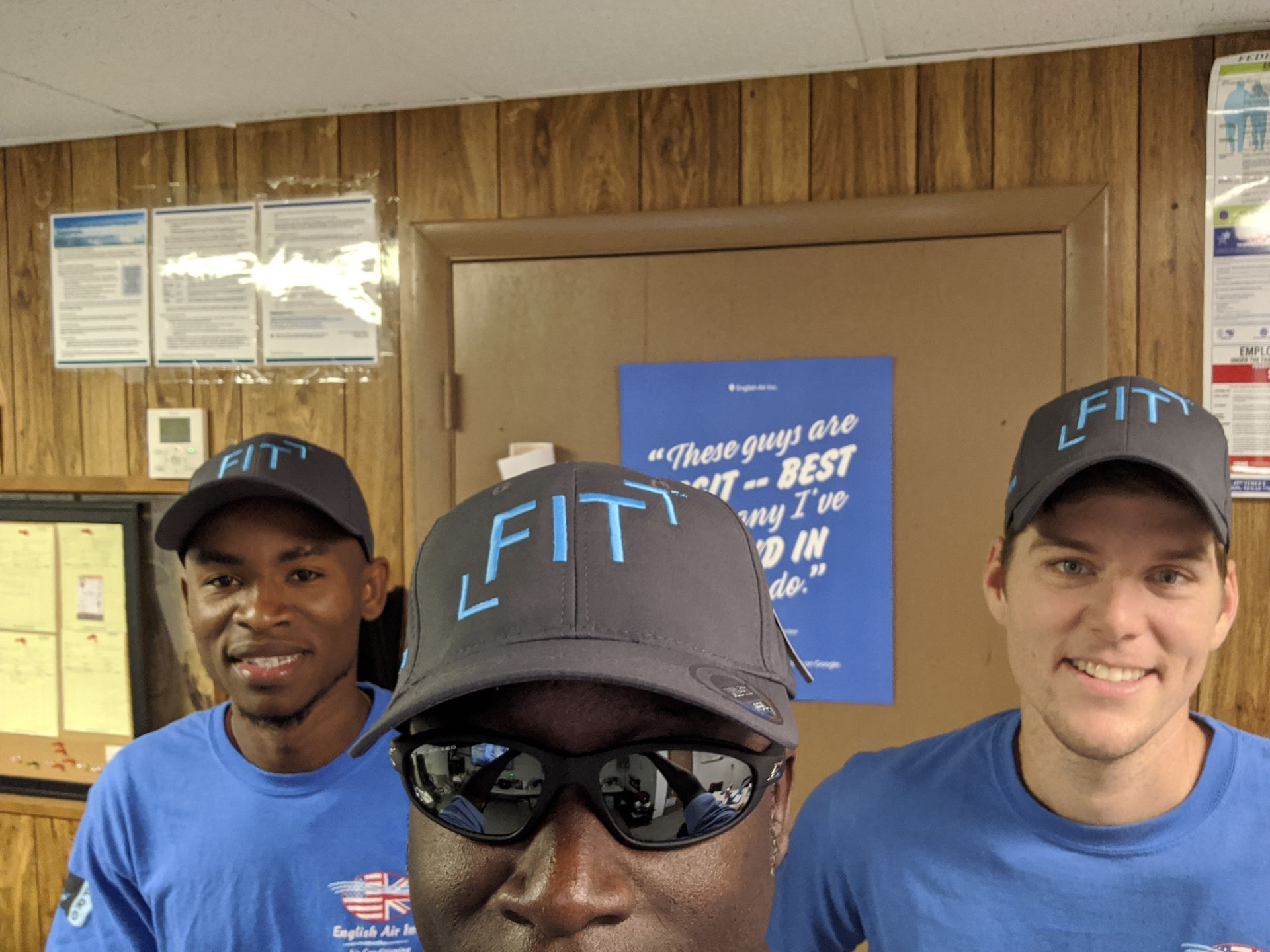 Three people in blue shirts and FIT hats posing indoors in front of a bulletin board