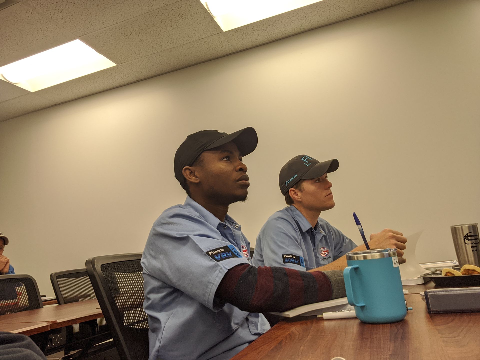 Two coworkers in blue uniforms sit at a table in a break room, one holding a pen beside a teal mug.
