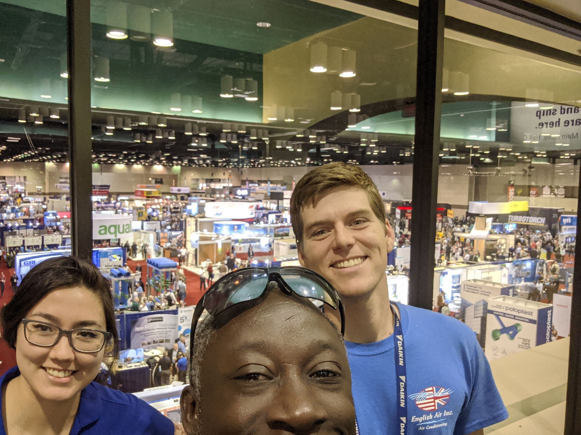 Three smiling people posing at a crowded indoor convention or trade show.