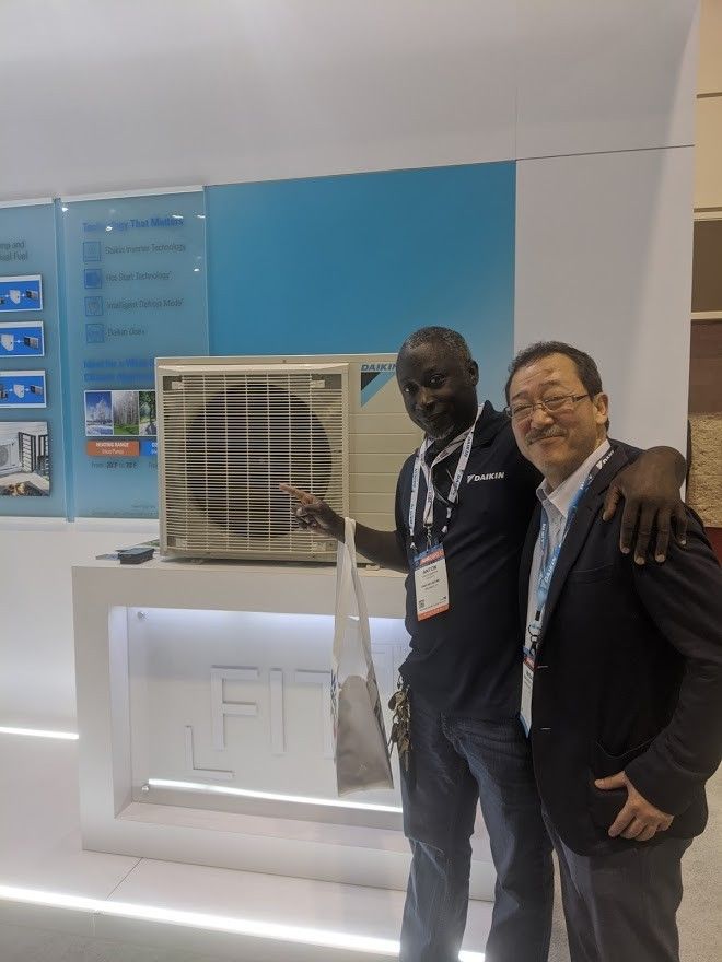 Two men standing beside a display booth and window air conditioner, smiling at the camera.