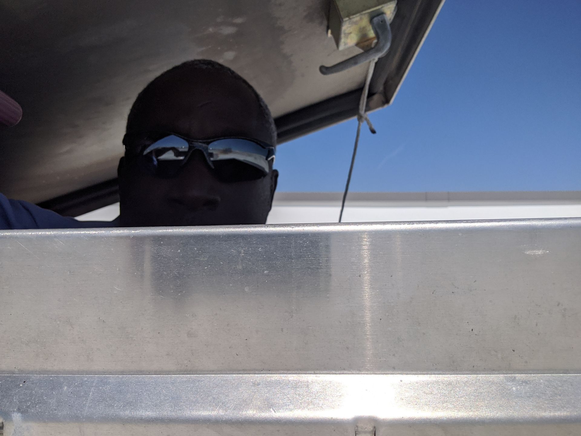 Person wearing sunglasses peeks over a silver car door under a bright blue sky
