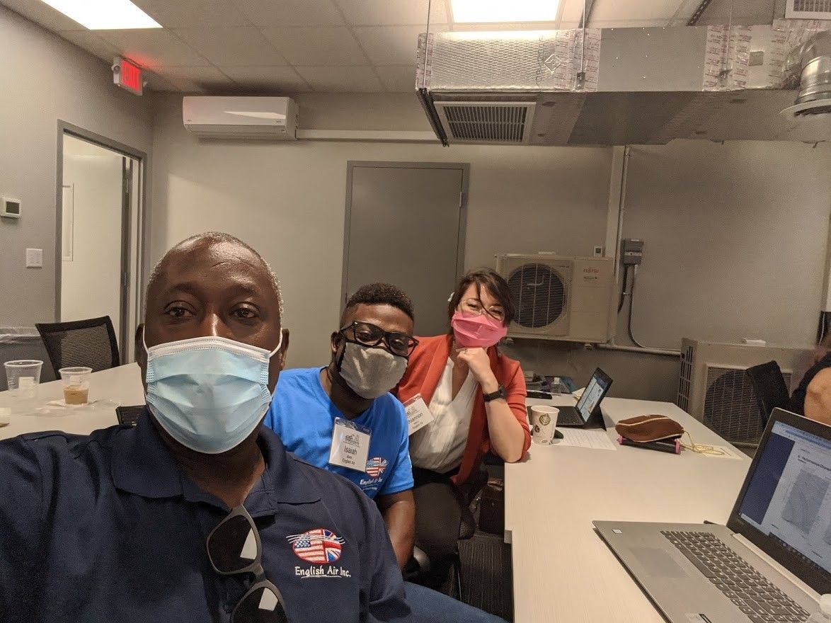 Three masked coworkers posing in an office break room, one at a laptop in the foreground