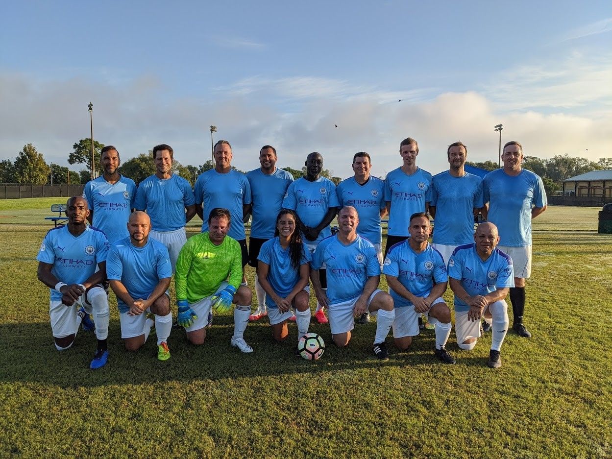 Soccer team posing on a grassy field, wearing blue and white uniforms with a bright green goalkeeper jersey.