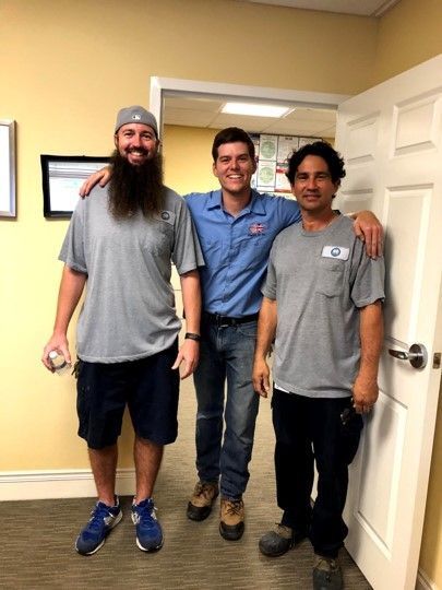 Three men smiling in a doorway, wearing casual shirts and jeans in a hallway.