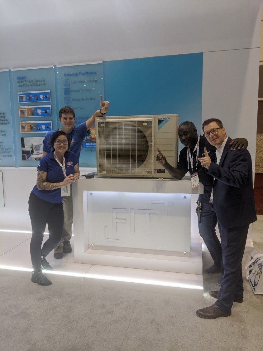 Four people pose around a large air conditioner display at a trade show booth.
