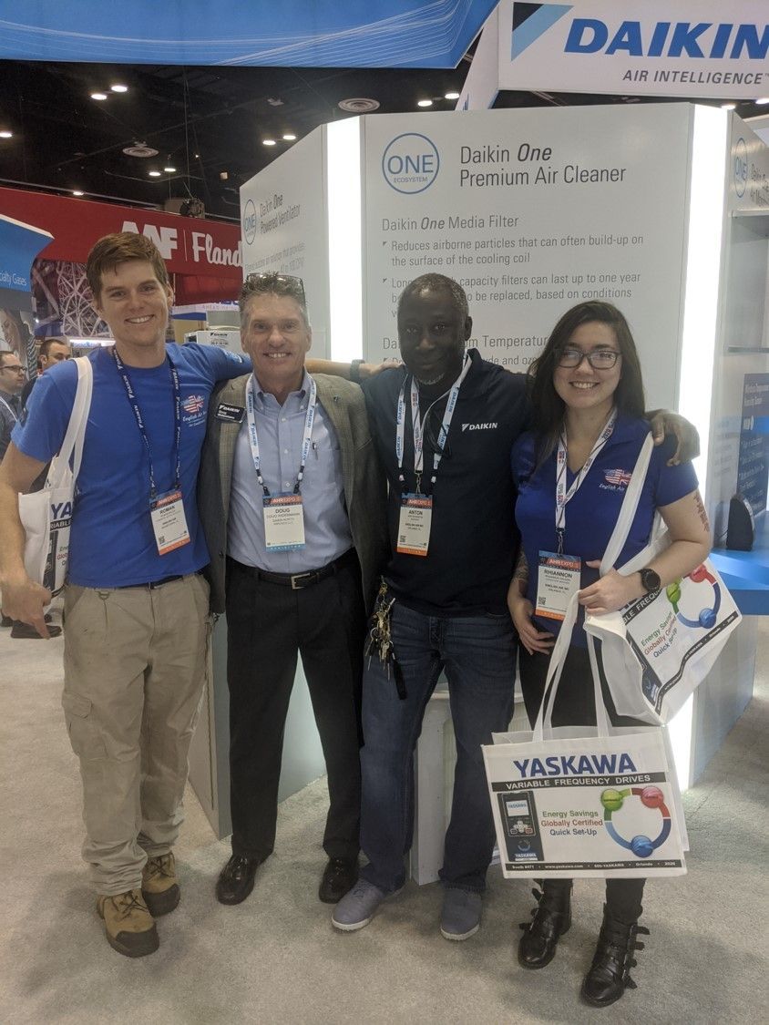 Four people posing at a Daikin trade show booth, holding branded bags and wearing conference lanyards.