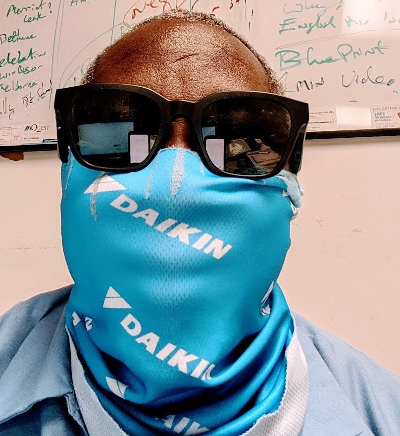 Person wearing black sunglasses and a blue Daikin face covering in front of a whiteboard.