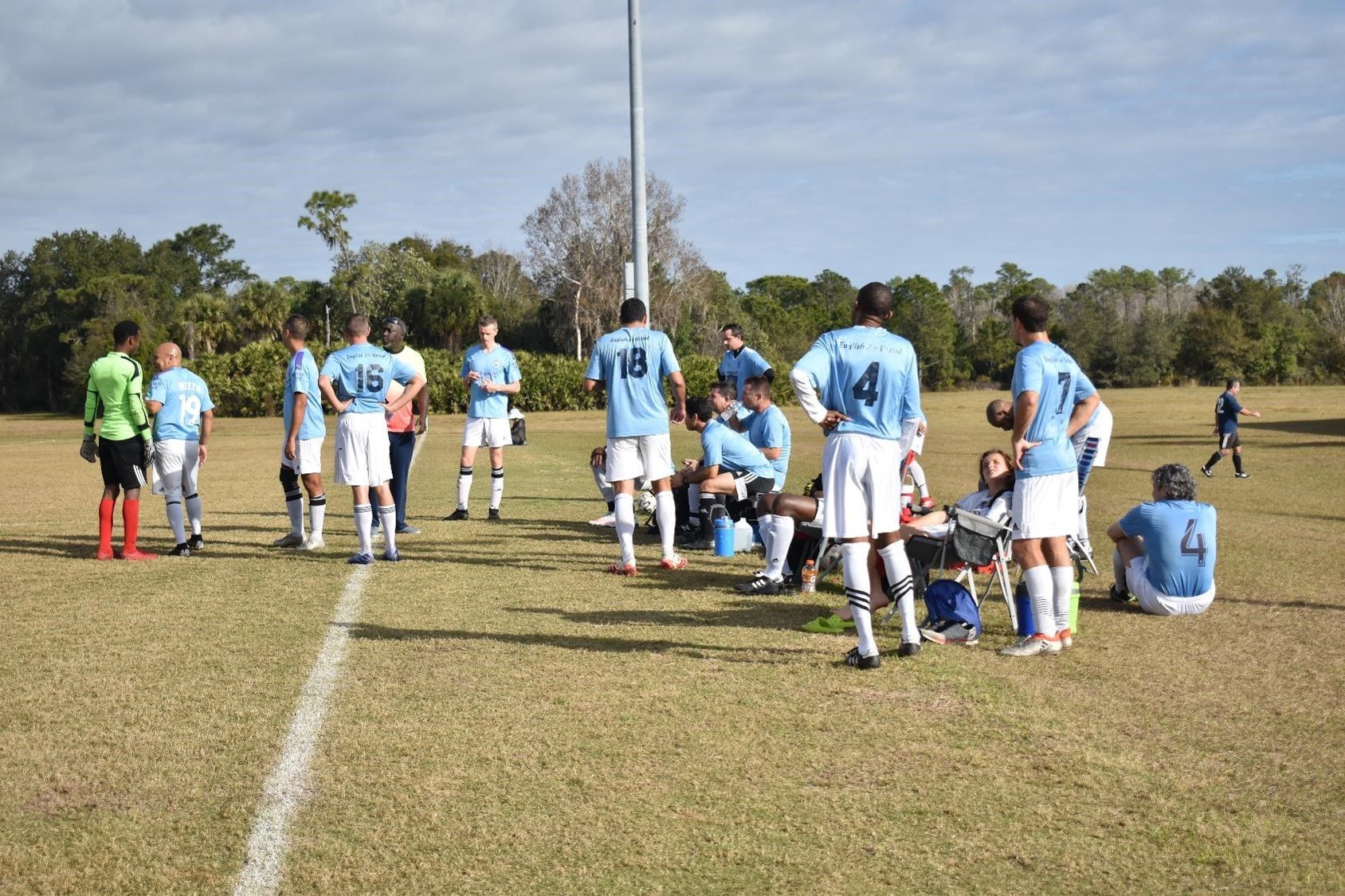 Soccer players in light blue jerseys huddle on a grassy field during a break in play
