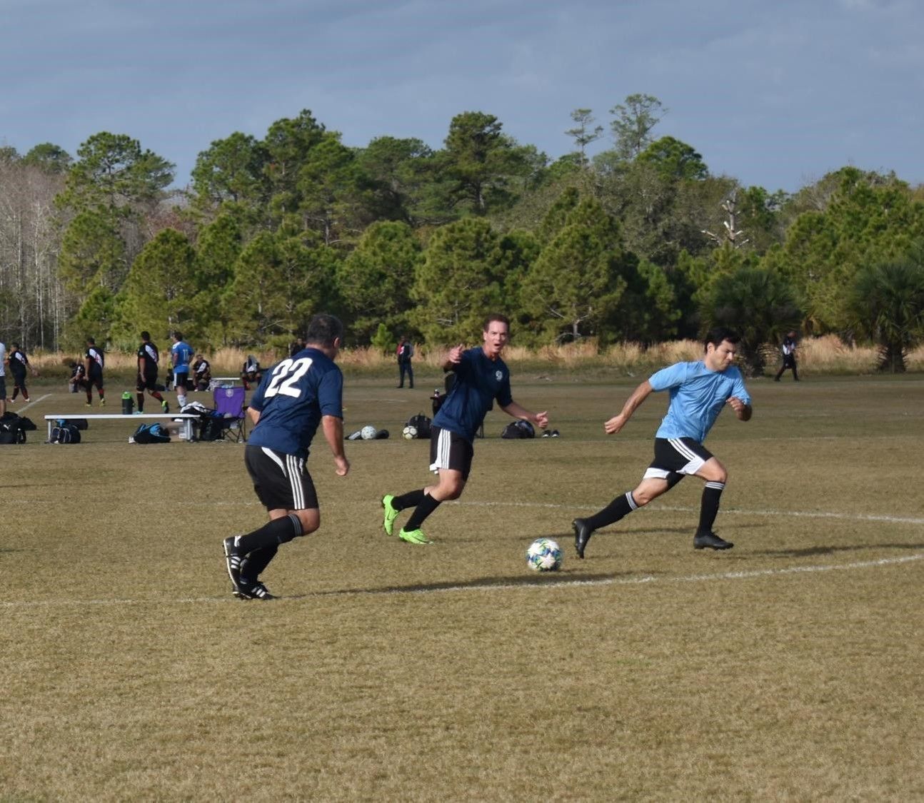 Soccer players in black and blue uniforms chase the ball on a grassy field with trees in the background