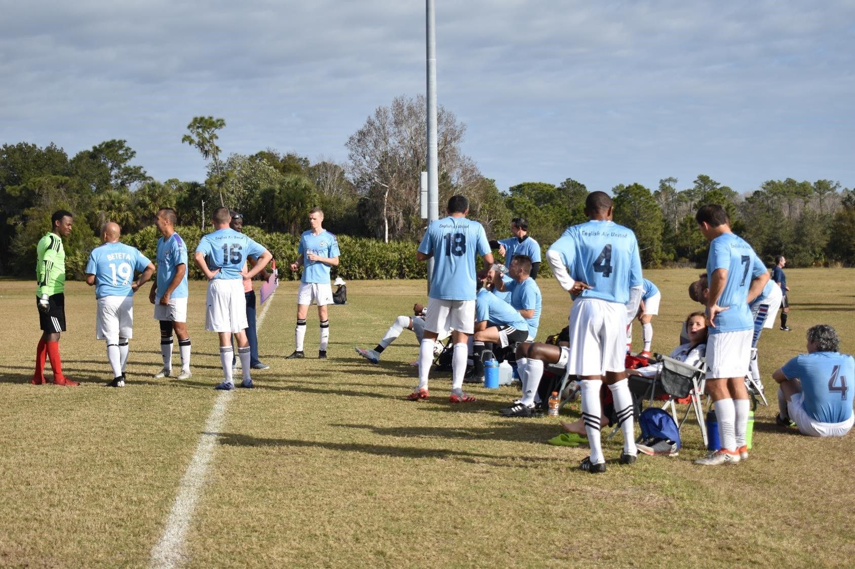 Soccer team in light blue jerseys huddles on a grassy field during a pause in play