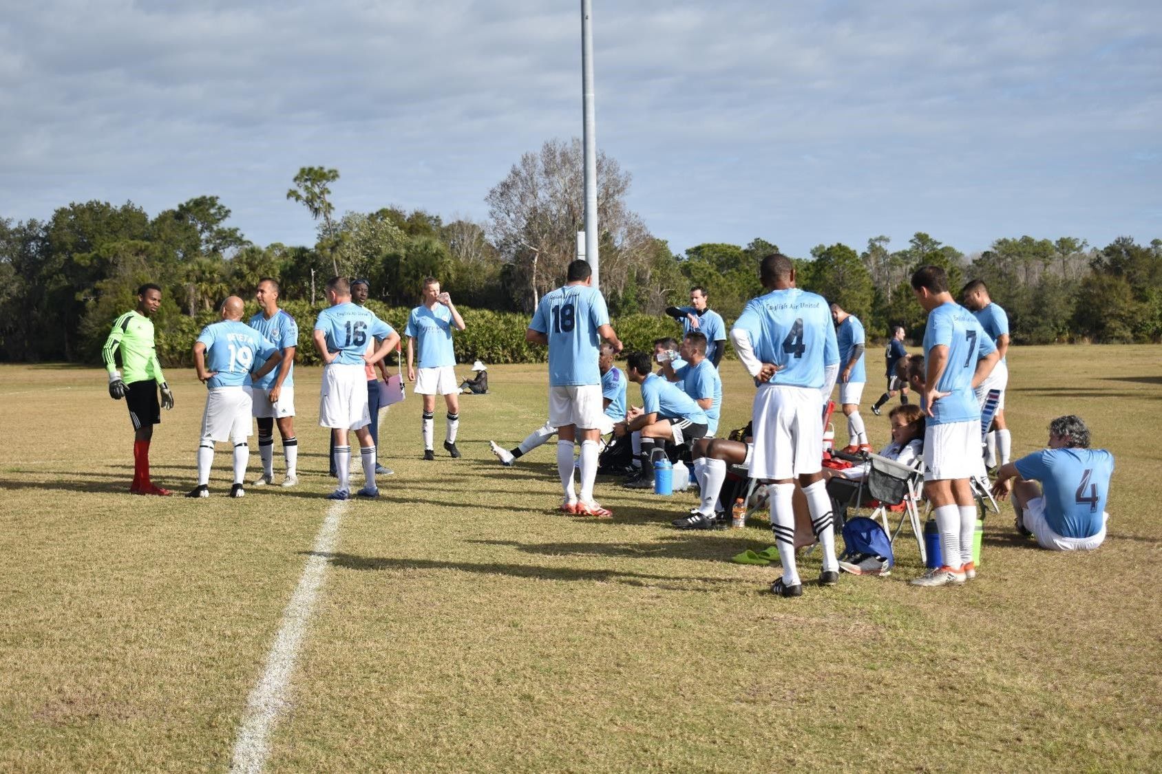 Soccer players in light blue uniforms huddle on a grassy field during a break in play