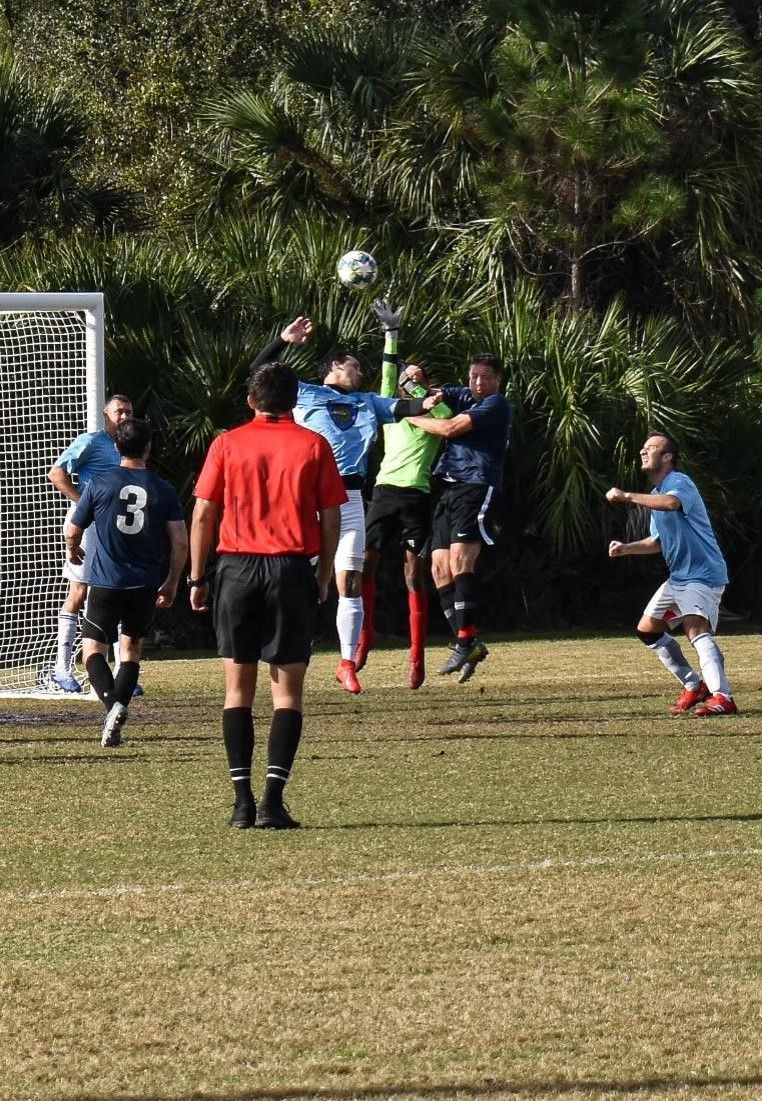 Soccer players contest a high ball near the goal as the referee watches.