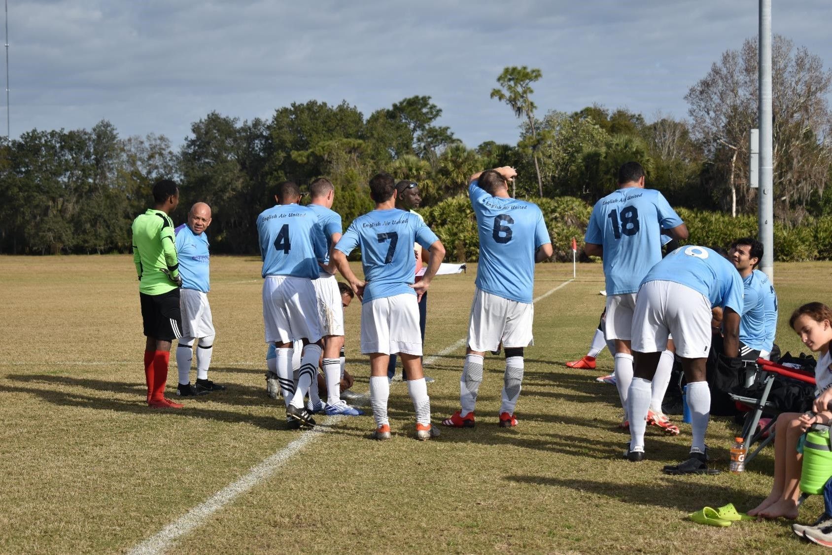 Soccer team in light blue jerseys huddles on a grassy field during a game break
