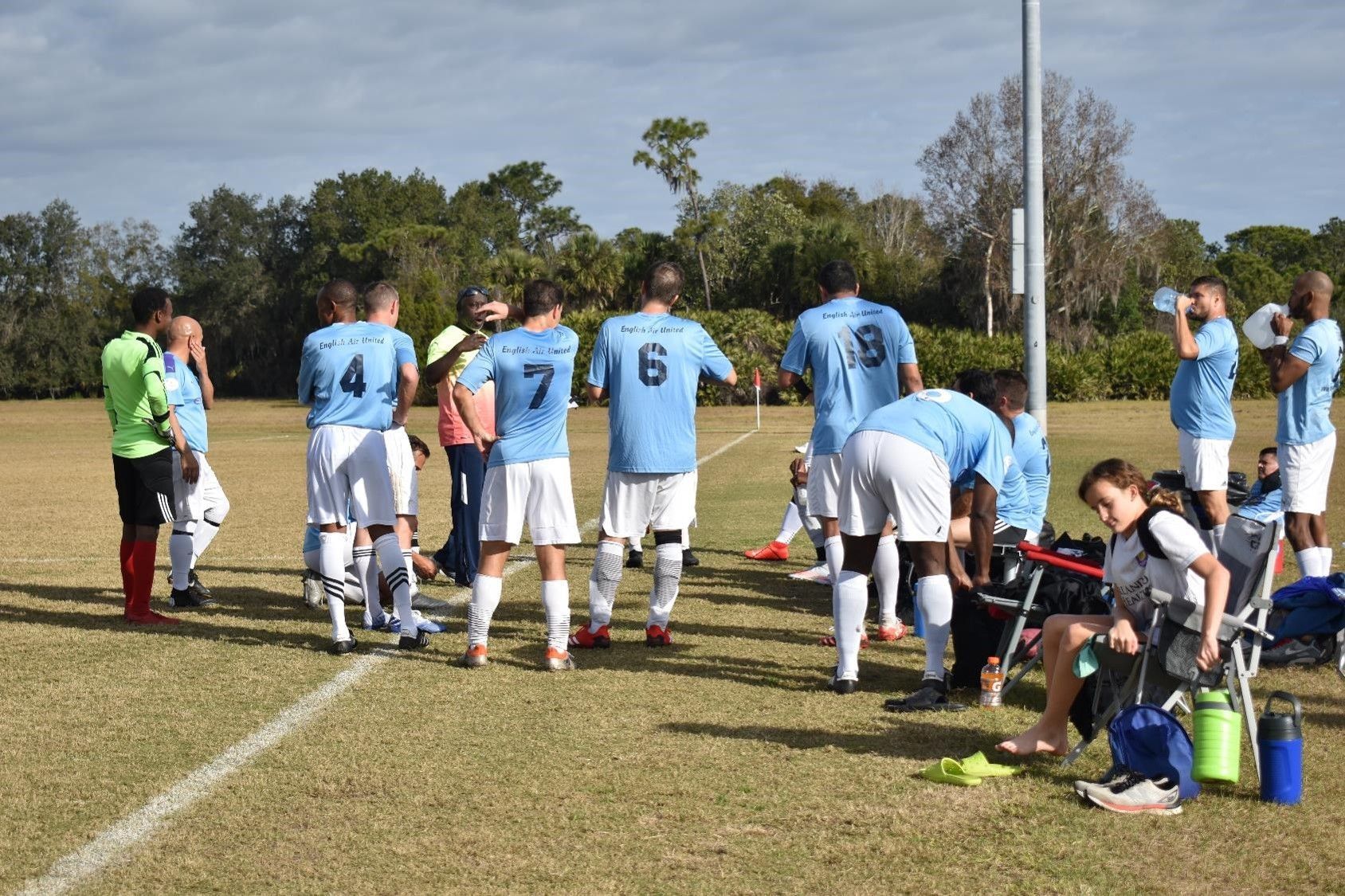 Soccer team in light blue jerseys huddles on a field beside the bench during a break.