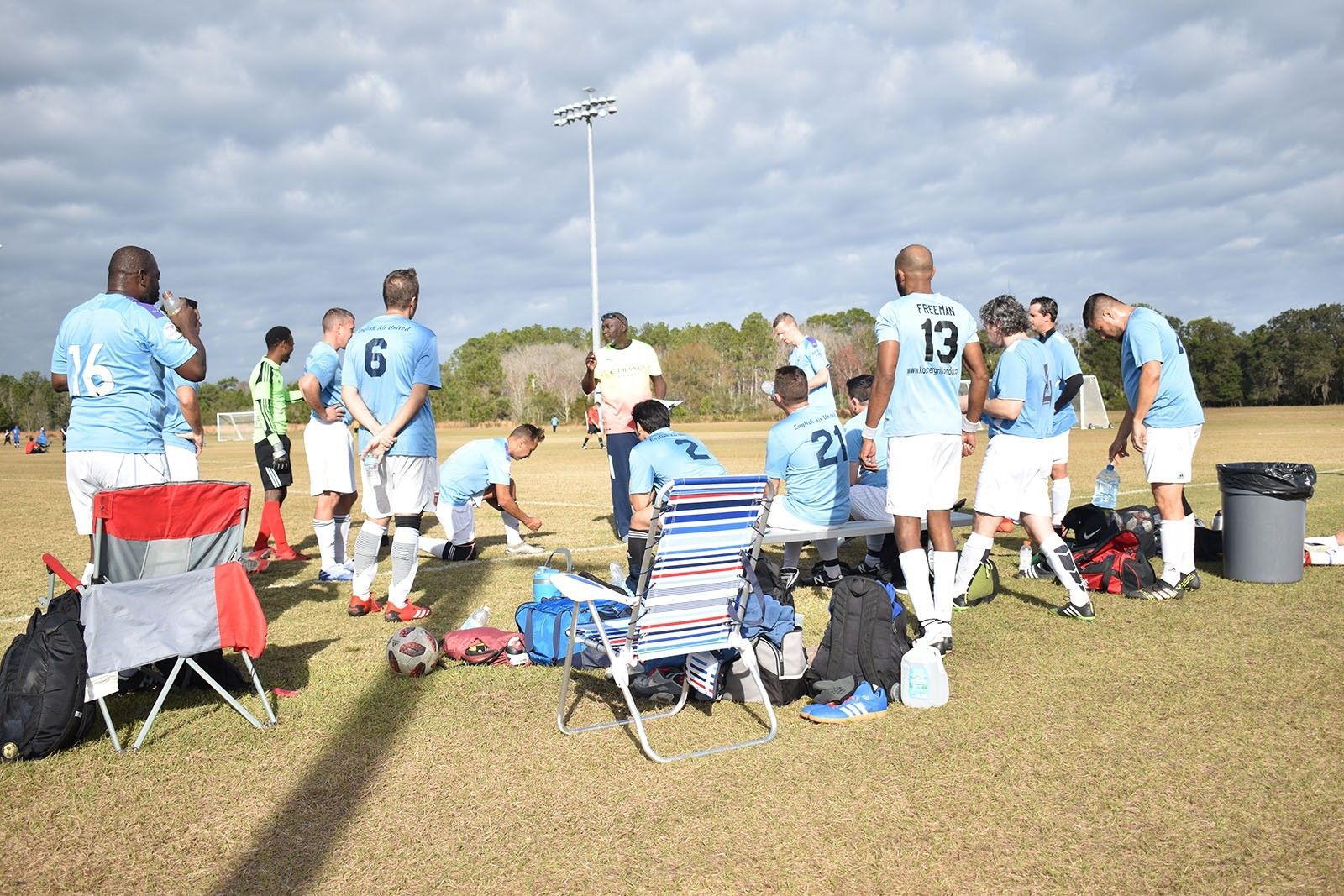 Soccer players in light blue jerseys huddle on a grassy field beside folding chairs and bags.