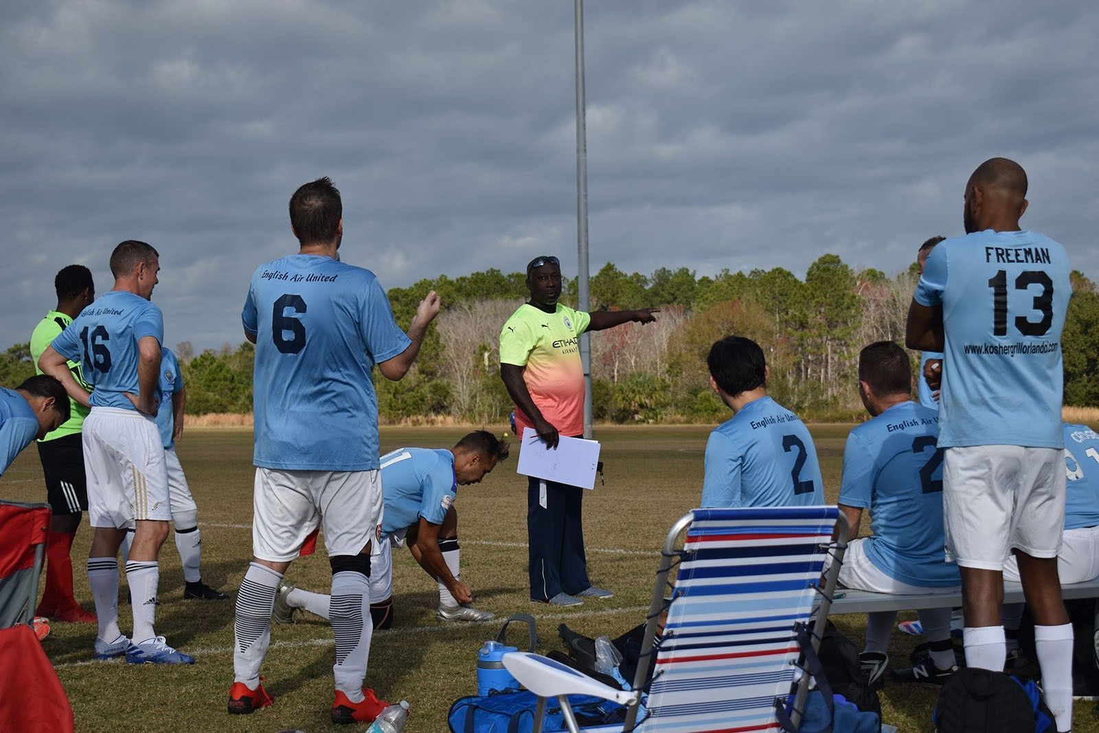 Soccer players in blue jerseys huddle on the sideline, listening to a coach near a grassy field