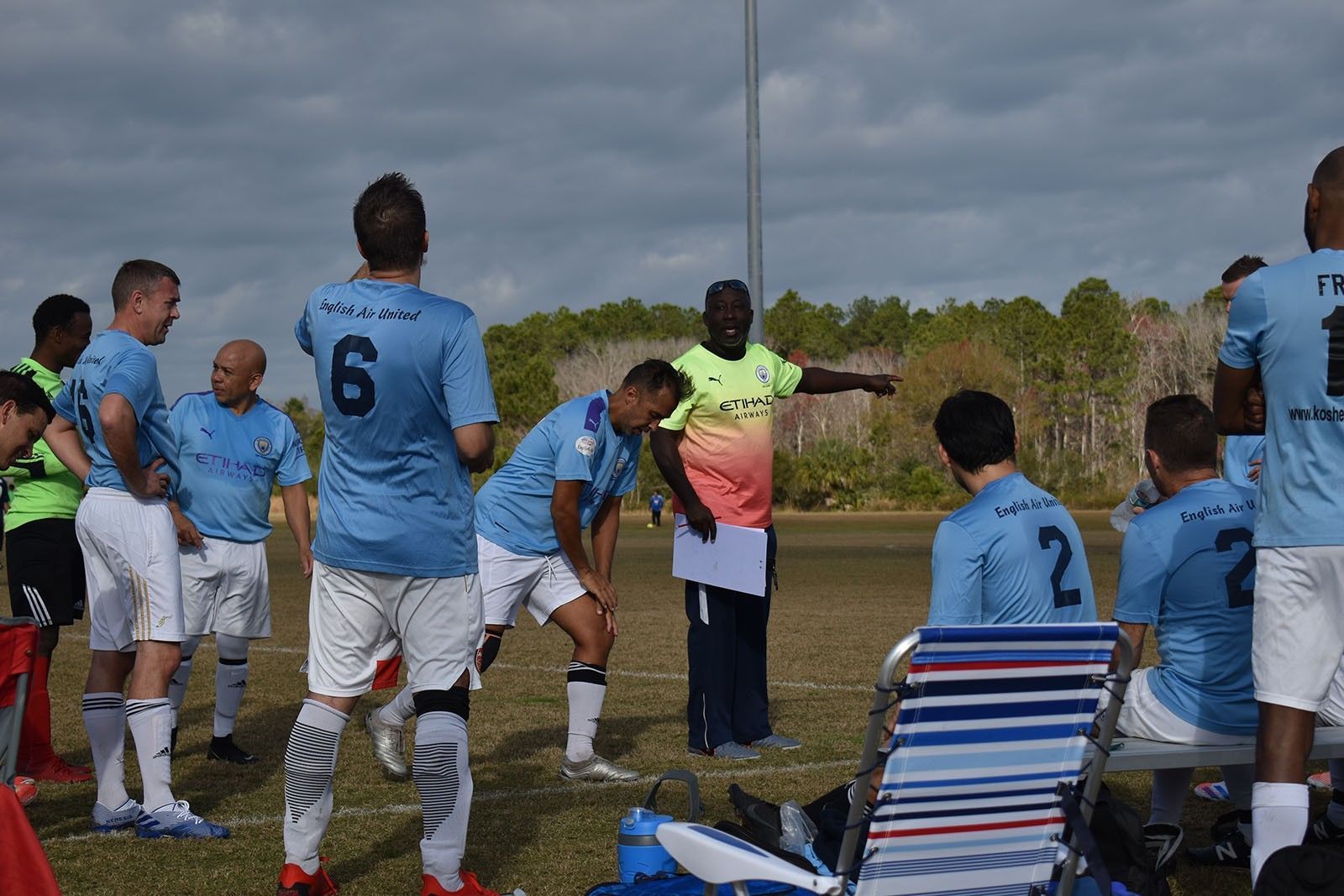 Soccer team huddled on a sideline under cloudy skies, with players in blue jerseys and a coach speaking.