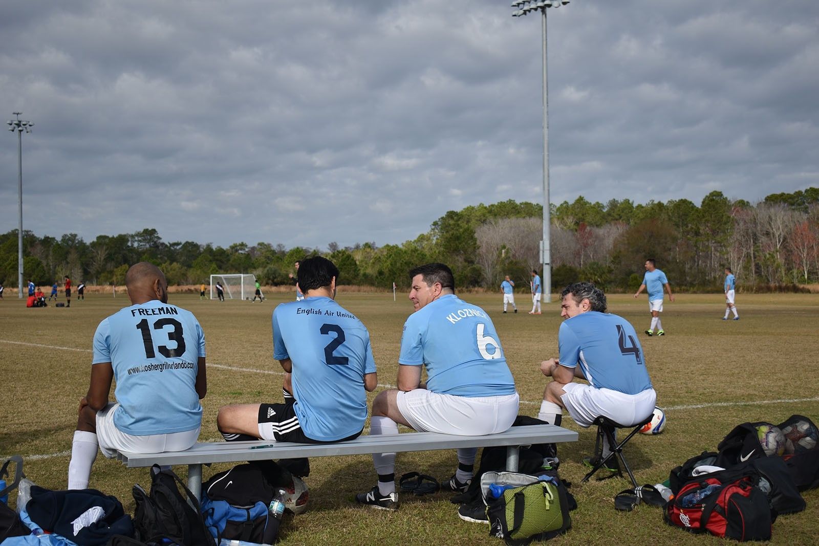 Soccer players in blue jerseys sit on a bench during a game on a grassy field