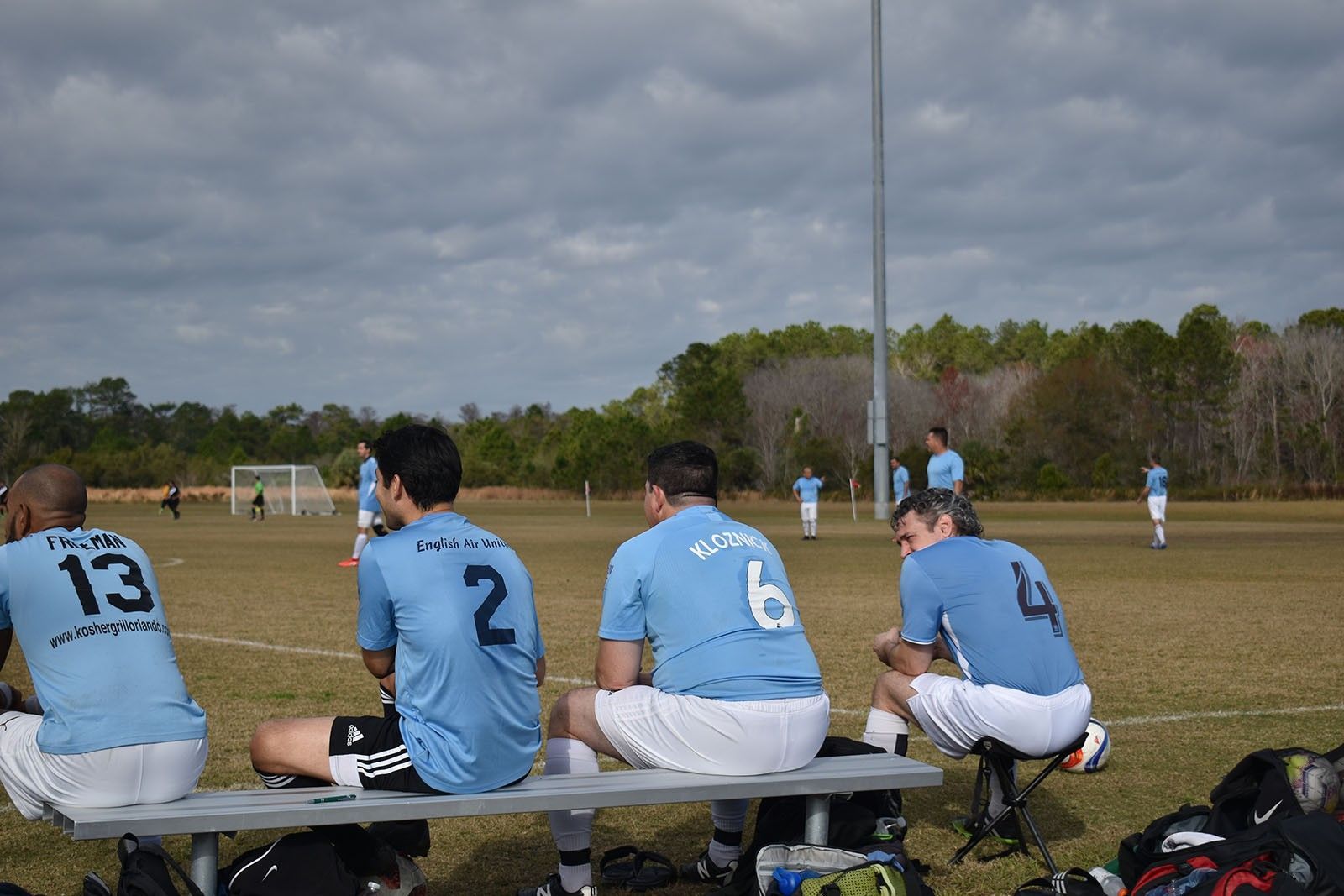 Soccer players in blue jerseys sit on a bench watching a game on a cloudy field