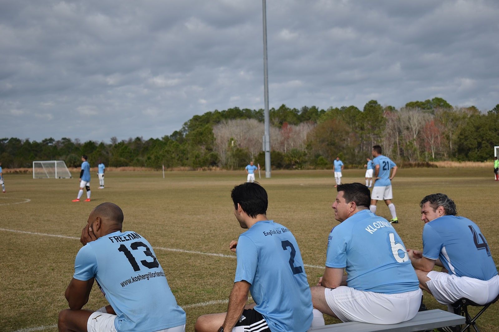 Soccer players in light blue jerseys sit on the sideline during a game on a grassy field.