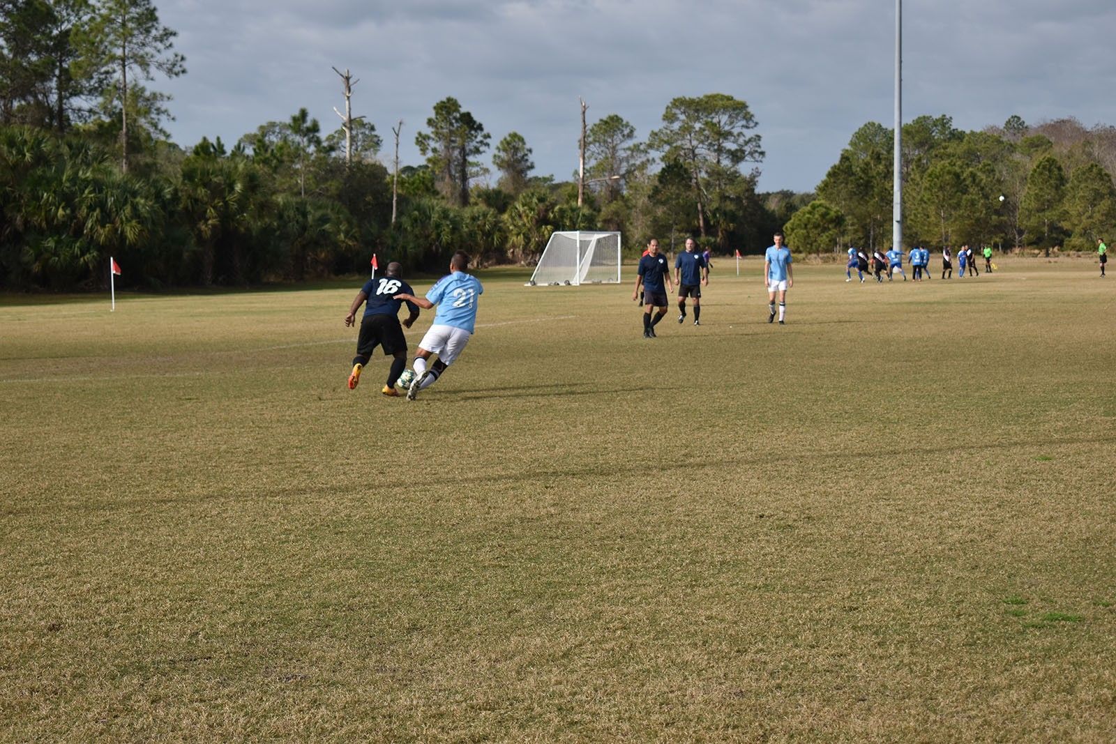 Soccer players chasing the ball on a grassy field near a goal under a clear sky