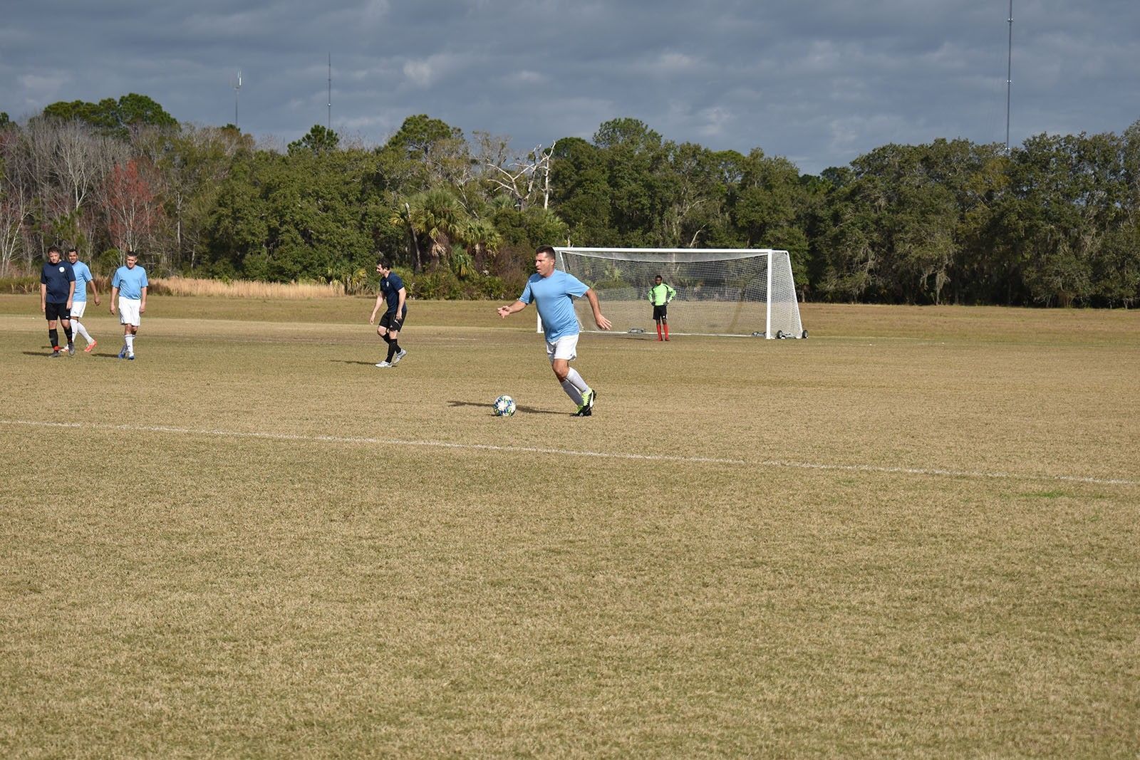 Soccer player in light blue kicks a ball toward a goal on a grassy field, with teammates nearby.