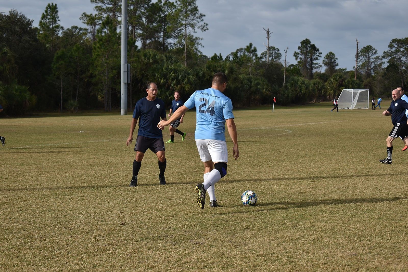 Soccer players on a grassy field, one in blue dribbling toward the goal, others in dark uniforms nearby