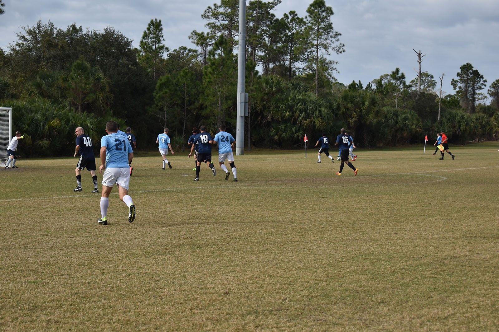 Soccer players in light blue and dark uniforms play on a grassy field with trees in the background