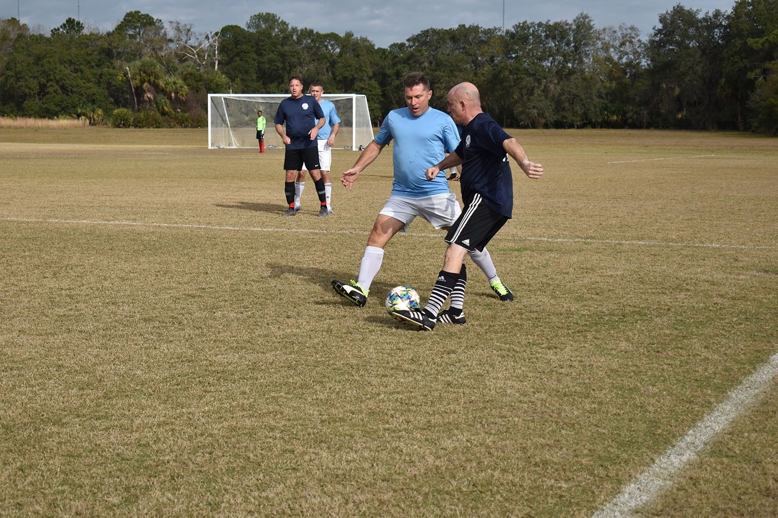 Two soccer players battling for the ball on a grassy field, with a goal in the background.