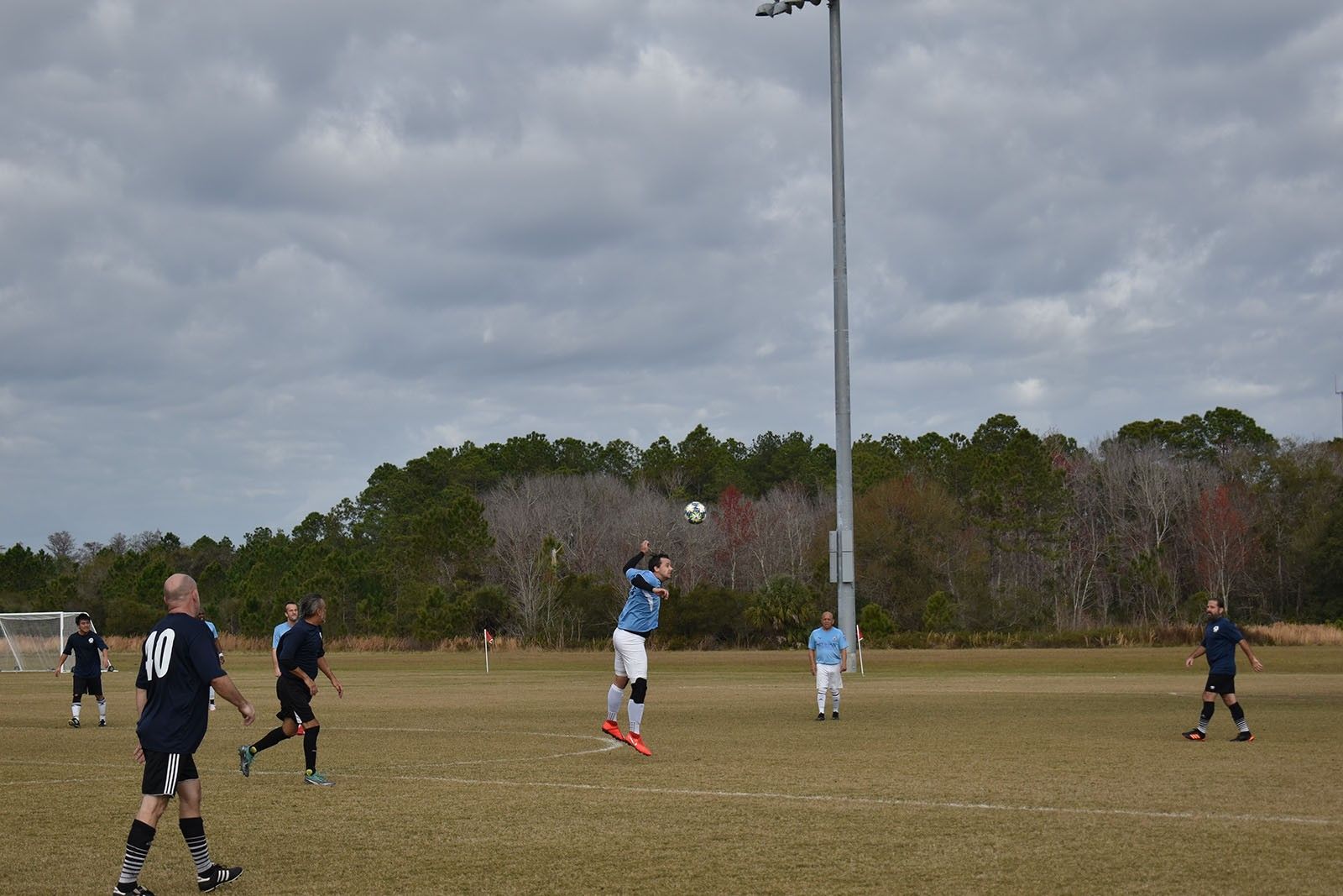 Soccer players on a grassy field under cloudy skies, with a tall light pole in the center.