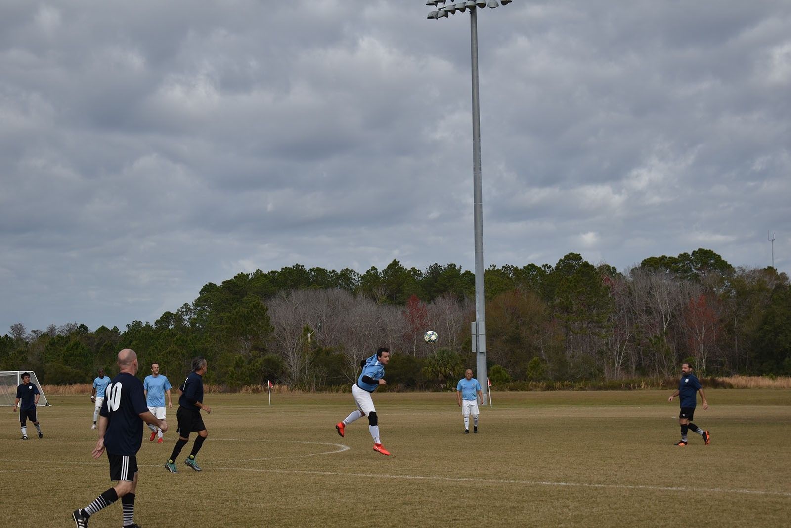 Soccer players on a grassy field under cloudy skies, with one player mid-kick near the center pole