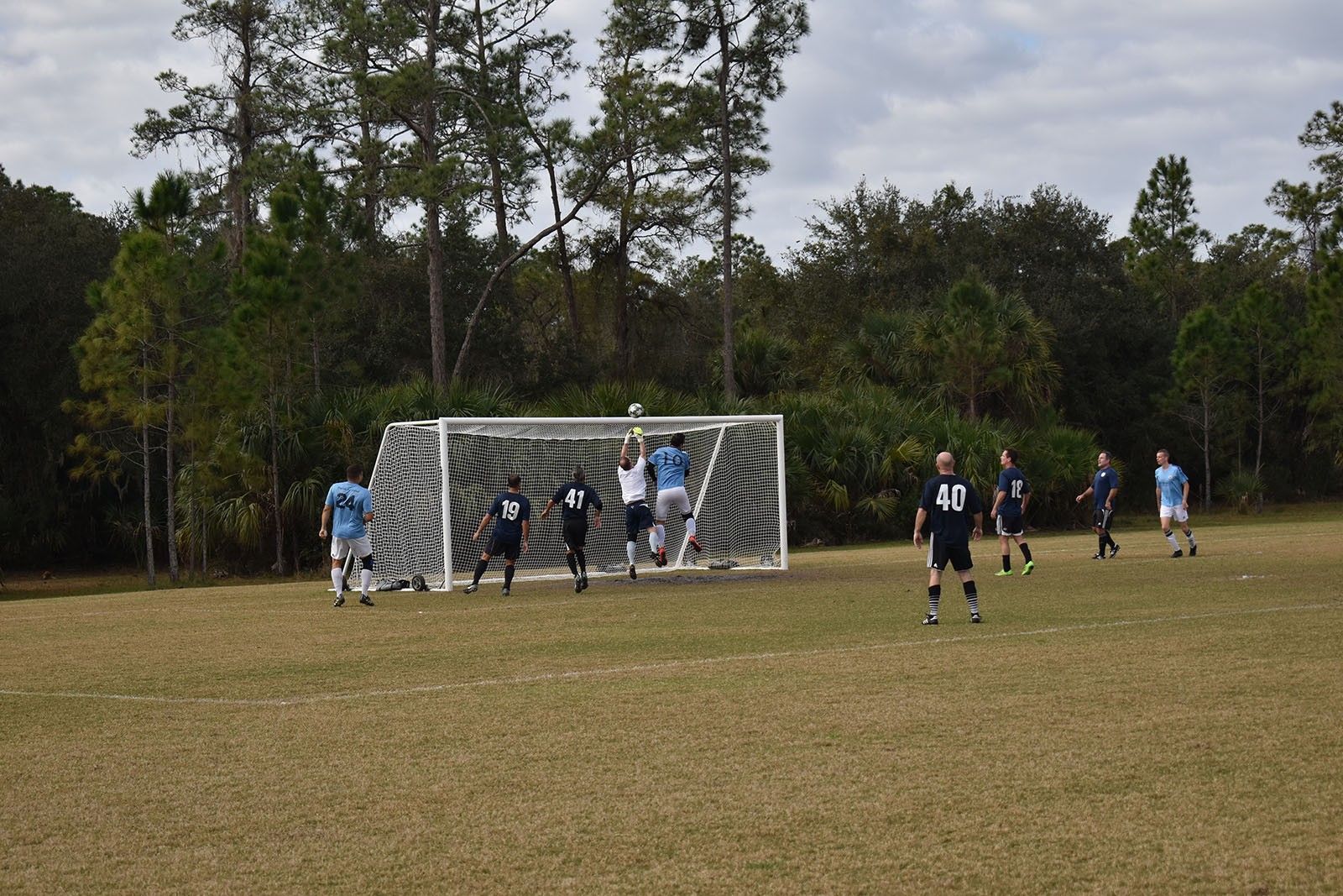 Soccer players near the goal on a grassy field, with spectators and trees in the background