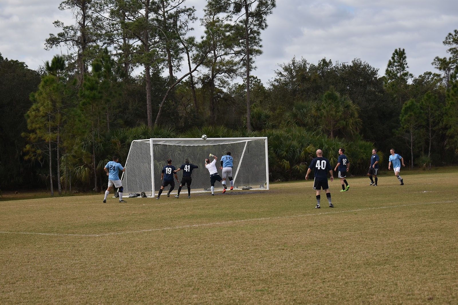 Soccer players crowd the goal as a shot is attempted on a grassy field