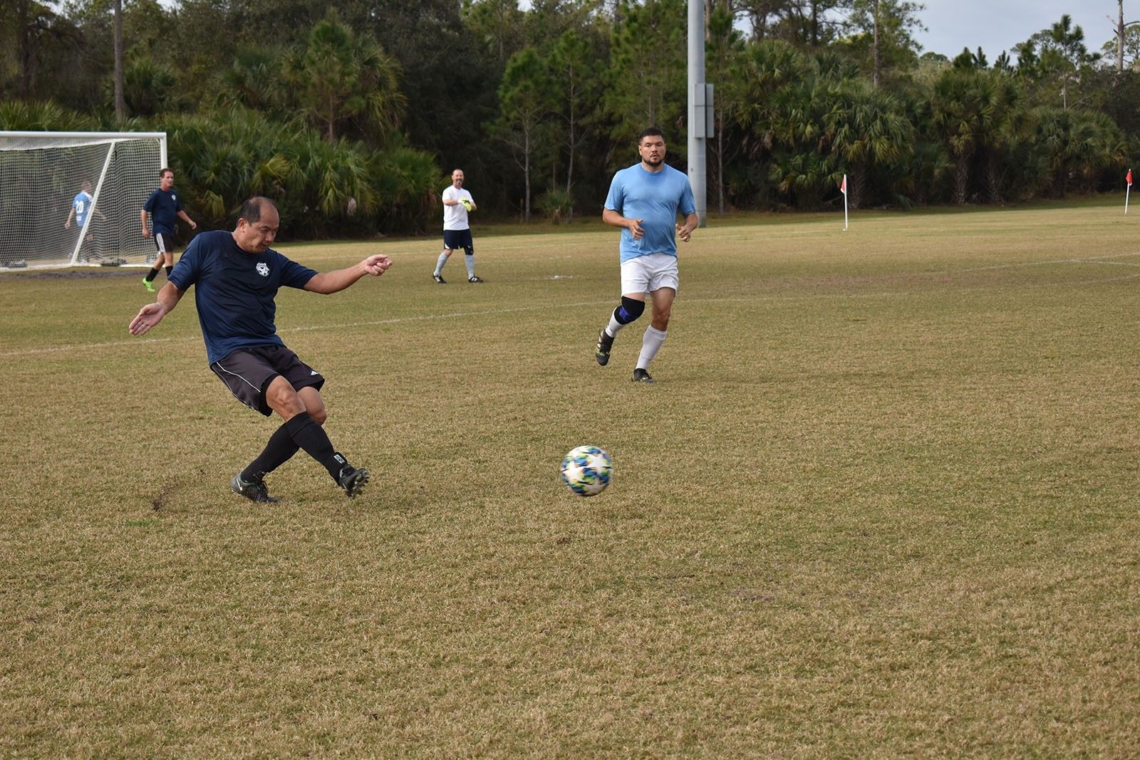 Soccer players chasing a ball on a grassy field near a goal.