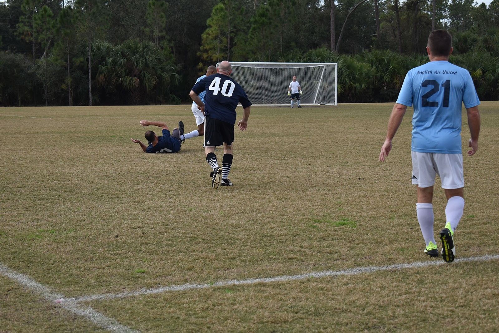 Soccer players on a grassy field near the goal, one player fallen, another in black running toward the ball
