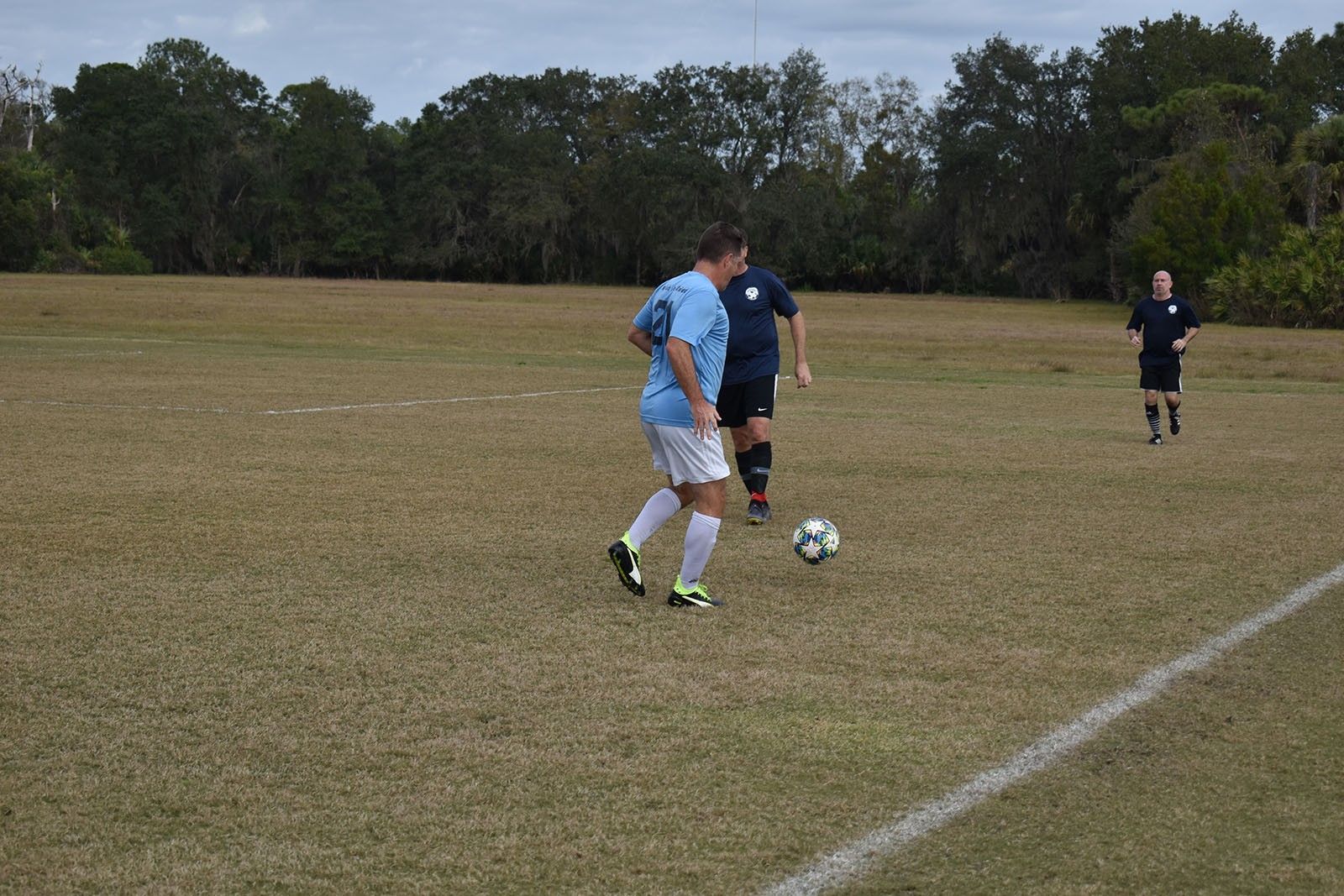 Two soccer players contest the ball on a grassy field, with a referee and trees in the background.