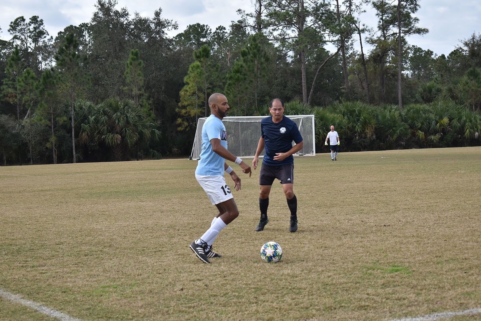 Two soccer players contest the ball on a grassy field with a goal and trees in the background.