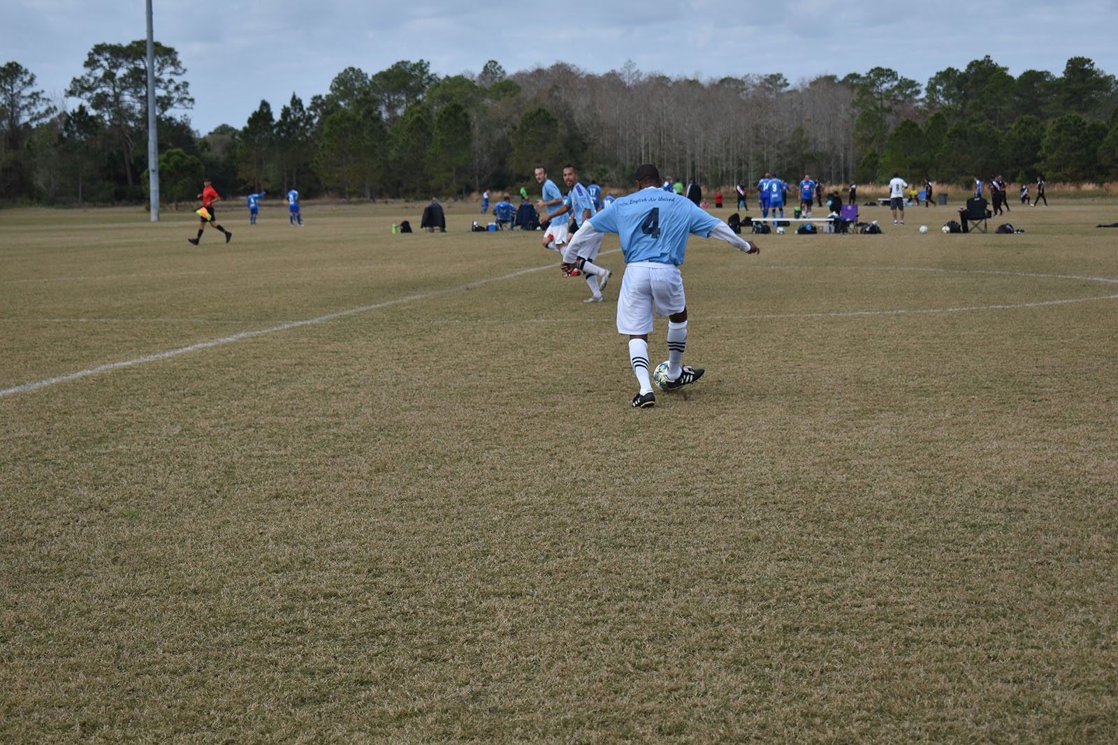 Youth soccer players on a grassy field, with one in a light blue jersey dribbling the ball away from the camera.