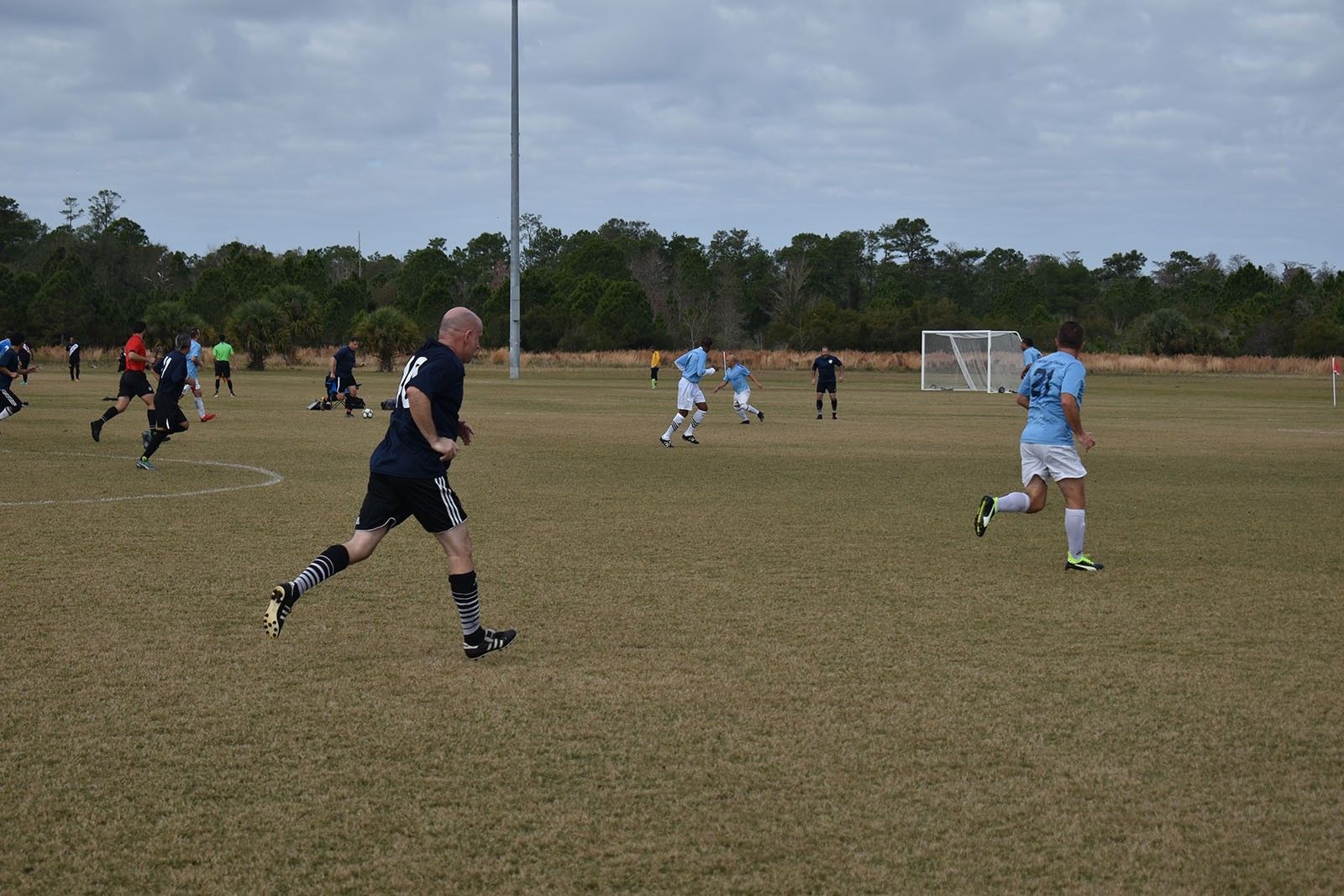 People playing soccer on a grassy field, with a goal in the background under an overcast sky.