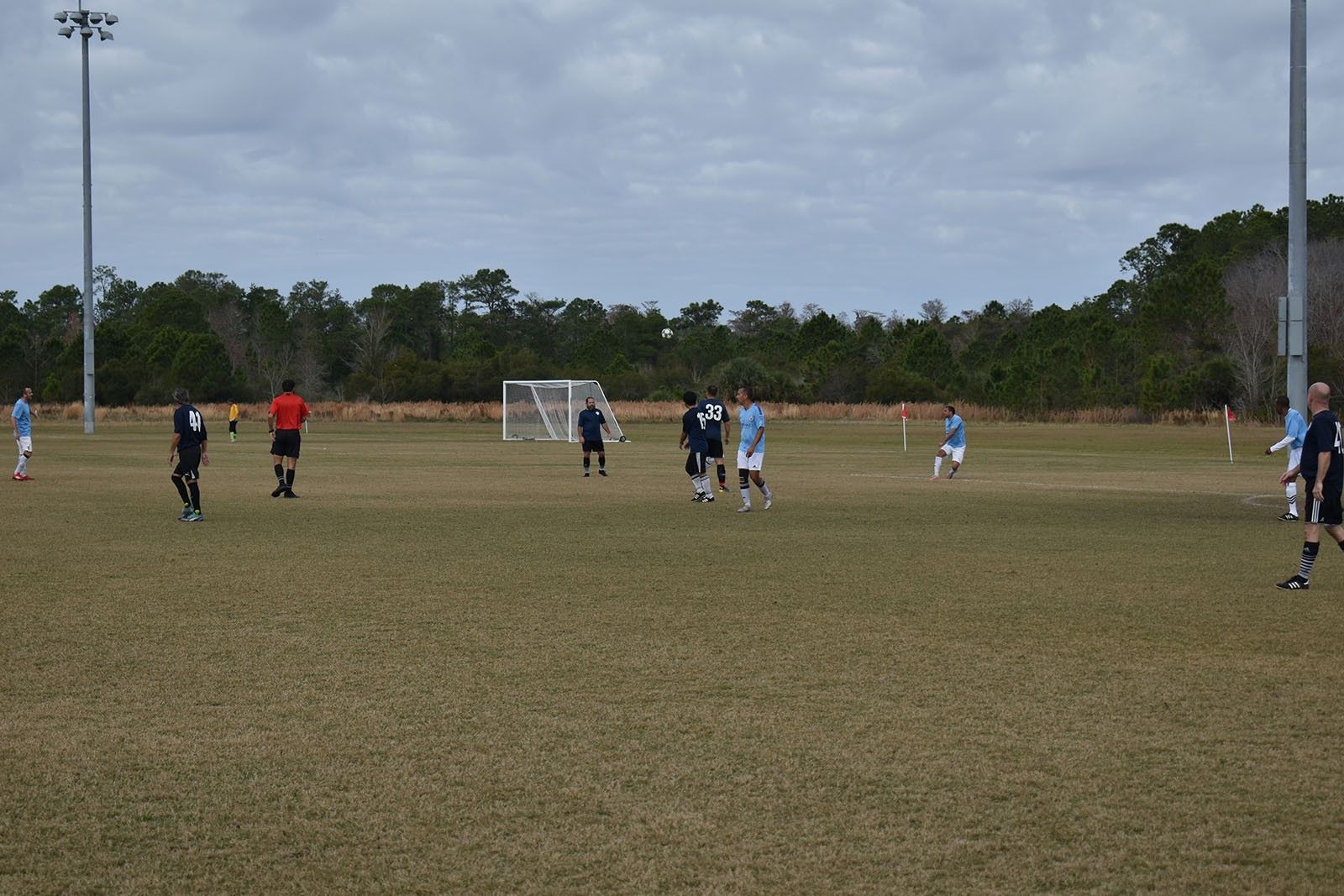 Soccer players on a field near a goal, with a referee and tall light poles under a cloudy sky