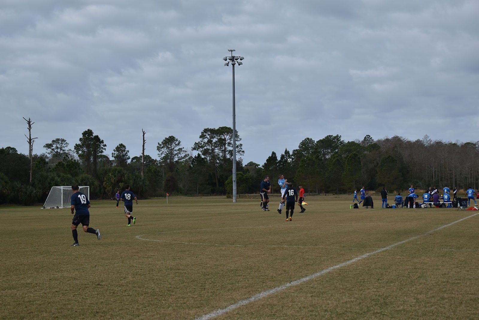 Soccer players on a grassy field under cloudy skies, with a group gathered near the sideline.