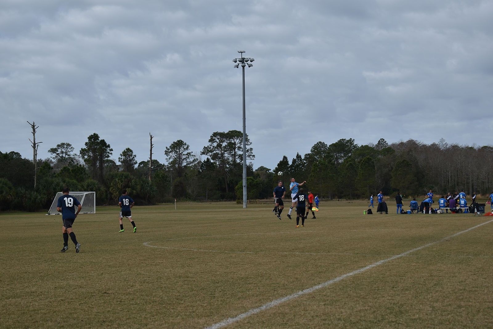 Soccer players on a grassy field under cloudy skies, with one group near the sideline and a tall light pole in the center.